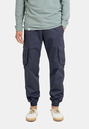 Mann trägt marineblaue Cargo-Jogginghosen mit elastischen Bündchen, ein hellgrünes Langarmshirt und weiße Sneakers mit beigen Schnürsenkeln.