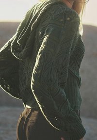 Pull en tricot vert foncé avec un motif de feuilles texturé, doté d'une capuche et d'un ourlet côtelé, associé à un jean noir ajusté.