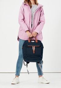 Granatowy plecak z brązowymi skórzanymi akcentami, z przednią klapą, paskami w paski i teksturowanym materiałem; zestawiony z różową kurtką i niebieskimi dżinsami.