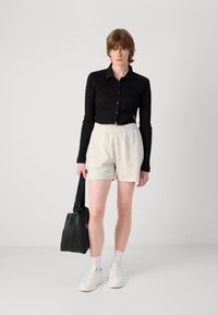 Svart, ribbet langermet skjorte med knapper, beige elastiske shorts med høyt liv, hvite joggesko og en svart skulderveske. Teksturerte stoffdetaljer er synlige.
