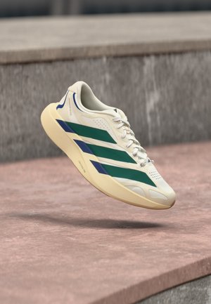 Chaussure de sport blanche avec des rayures vertes et bleues suspendue au-dessus d'une surface en pierre et d'un mur de pierre gris en arrière-plan.