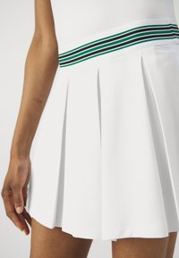 Λευκή πλισέ φούστα με αντίθετη ζώνη που διαθέτει πράσινες και μαύρες ρίγες. Λείο ύφασμα και ραμμένη εφαρμογή, αναδεικνύοντας λεπτομέρεια στο στρίφωμα.