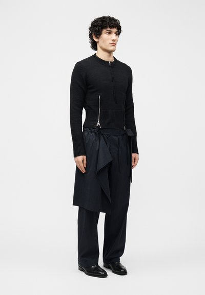 Άνδρας με σγουρά μαλλιά που φοράει μαύρο πουλόβερ με φερμουάρ και σκουρόχρωμο παντελόνι με ρίγες και ασύμετρο draped πάνελ, στέκεται μπροστά σε απλό φόντο.