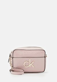 Jasnoróżowa torebka Calvin Klein na ramię z logo "ck" w kolorze złotym, czarną lamówką i odpinanym paskiem na nadgarstek na białym tle.