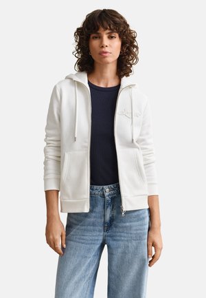 Woman with curly hair wearing a white zip-up hoodie over a black shirt and light blue jeans, standing against a plain background.
