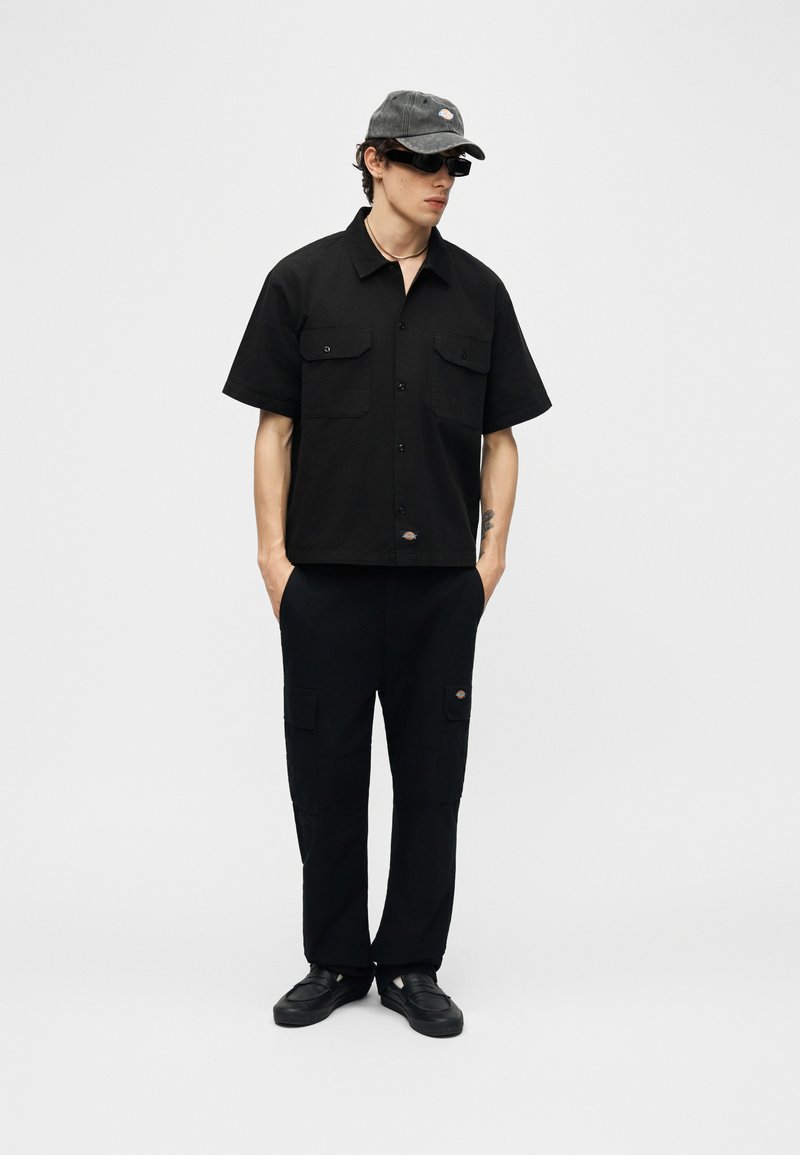 Giovane uomo con camicia nera a maniche corte con bottoni, pantaloni neri con tasche cargo, sneakers nere, cappellino grigio e occhiali da sole neri, con le mani nelle tasche.