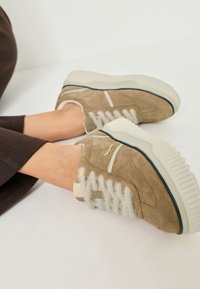 Tan ruskinds sneakers med hvide teksturerede snørebånd og en tykk, cremefarvet sål. Har et diskret guldlogo på siden.