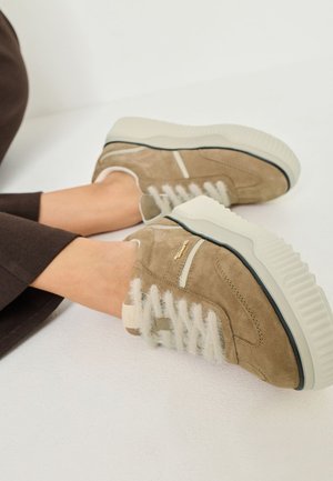 Beżowe zamszowe sneakersy z białymi teksturowanymi sznurówkami i grubą kremową podeszwą. Cechują się subtelnym złotym logo z boku.