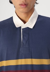 Marineblå polo skjorte med lys krage, med horisontale striper i gult og burgunder. Laget av bomull med knappeplacket og broderte detaljer.