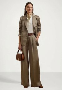 Γυναίκα φορώντας μπεζ σατέν κοστούμι με φαρδιά παντελόνια, λευκή μπλούζα, κρατώντας ένα καφέ τσάντα και φοράει χρυσά σκουλαρίκια.