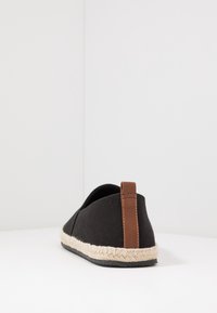 Black canvas shoe with a brown leather heel tab and woven jute midsole. Features a rounded shape and rubber outsole for grip.
