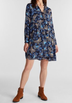 Kobieta w granatowej sukience w kwiaty z długim rękawem i dekoltem w kształcie litery V, nosząca brązowe zamszowe botki, stojąca na jasnym, jednolitym tle.