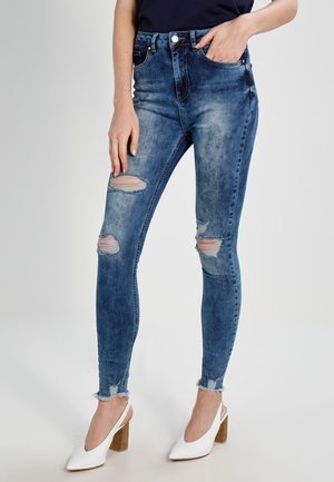 Kvinde iført blå revnede skinny jeans med frynsete kanter og hvide blokhælede slingback-sko, hånd hviler i lommen.