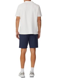 Herren Poloshirt mit kurzen Ärmeln in Weiß, kombiniert mit Marineblauen Shorts. Das Outfit umfasst graue Sneakers und weiße Knöchelsocken.