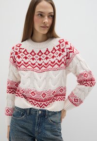 Maglione a maglia color crema con motivi geometrici rossi. Presenta una texture a trecce e scollo a costine. Abbinato a jeans blu scuro.