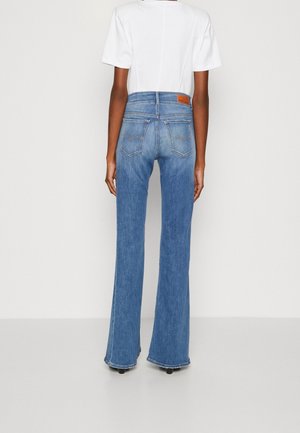 Persoon die een lichtblauwe jeans met hoge taille en achterzakken draagt, gecombineerd met een wit topje met korte mouwen, staand tegen een effen witte achtergrond.