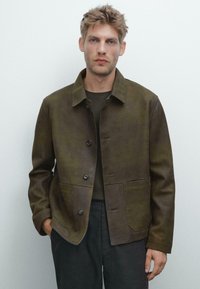 Braune Lederjacke mit Kragen, Knopfverschluss und zwei vorderen Taschen. Texturierte Oberfläche, taillierte Passform, über einem dunklen Hemd getragen.