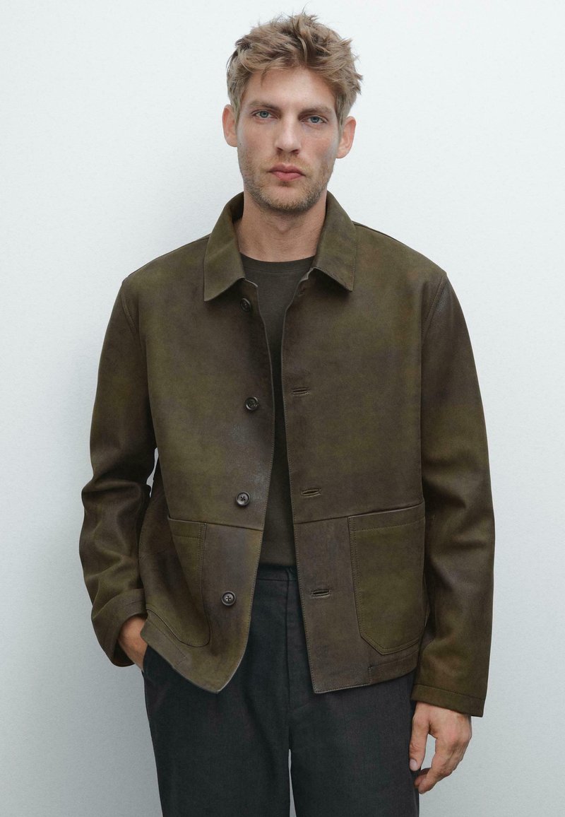 Braune Lederjacke mit Kragen, Knopfverschluss und zwei vorderen Taschen. Texturierte Oberfläche, taillierte Passform, über einem dunklen Hemd getragen.