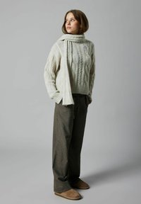 Pull en tricot torsadé couleur crème avec un col montant, associé à un pantalon à carreaux dans des tons de marron et de vert, et des chaussures slip-on beiges.