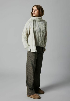 Pull en tricot torsadé couleur crème avec un col montant, associé à un pantalon à carreaux dans des tons de marron et de vert, et des chaussures slip-on beiges.