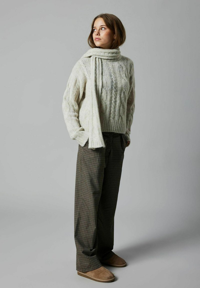 Pull en tricot torsadé couleur crème avec un col montant, associé à un pantalon à carreaux dans des tons de marron et de vert, et des chaussures slip-on beiges.