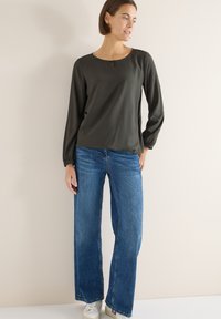 Donna in piedi contro una parete beige che indossa una blusa verde scuro a maniche lunghe, jeans larghi blu e sneakers bianche.