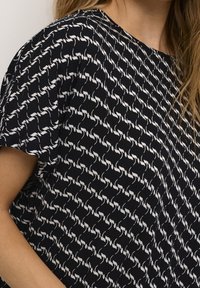 Gros plan d'une personne portant un haut noir avec un motif abstrait en forme de chaîne blanche répétée et des manches courtes.