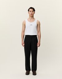 Λευκό μπλουζάκι χωρίς μανίκια με κεντημένο μαύρο λογότυπο συνδυασμένο με ψηλόμεσες μαύρες παντελόνες. Καφέ παπούτσια ολοκληρώνουν το σύνολο. Ουδέτερο φόντο.