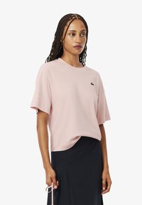 Camiseta de manga corta color rosa claro hecha de algodón con cuello redondo y detalle de logotipo bordado en el lado izquierdo del pecho. Textura suave.