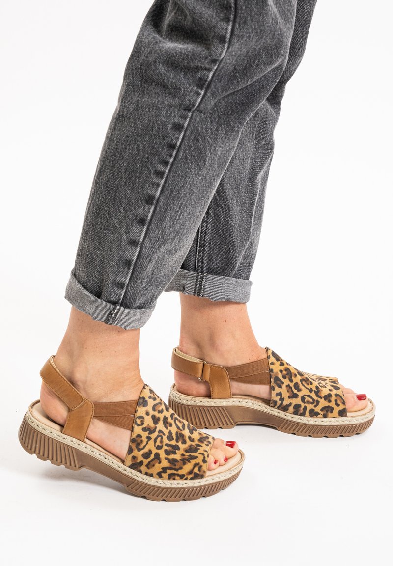 Leopard print sandals with tan accents, featuring a chunky sole, elastic straps, and an open design. Worn with rolled gray jeans.