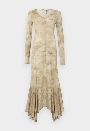 Moteris stovinti viena ranka ant klubų, dėvinti ilgą smėlio spalvos raštuotą suknelę su asimetriniu kraštu ir juodus lakuotus butus ant baltos fono.