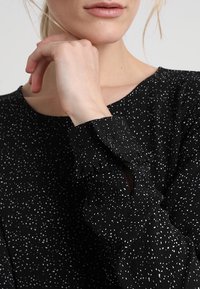 Blouse noire à motif tacheté blanc, avec un col rond et des poignets évasés bordés de volants. Tissu doux et texturé qui ajoute un intérêt visuel.