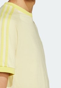 Ljusgul t-shirt med rund hals och korta ärmar, med tre vertikala ränder i en ljusare nyans av gult längs axlarna.