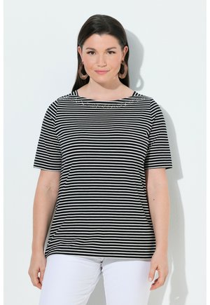 Kvinde med mørkt, glat hår iført sort-hvidt, vandret striped kortærmet skjorte, hvide bukser og store hoop øreringe, står op ad hvid væg.