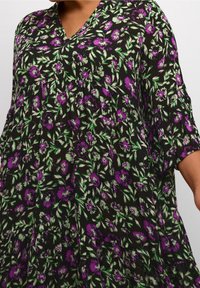 Schwarzes Kleid mit kurzen, weiten Ärmeln; präsentiert ein Blumendesign in Lila und Grün. V-Ausschnitt und lockere, fließende Form.