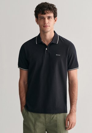 Marineblå polo-shirt med hvide piping ved kraven og ærmerne; har en tre-knaps knaplukning og et lille logo på brystet; lavet af bomuldsstof.