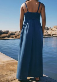 Robe longue bleue sans manches avec un corsage froncé, une jupe fluide et de fines bretelles. Portée près de l'eau, avec une texture lisse.