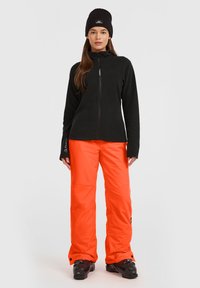 Svart fleece zip-up jacka, klara orange snöbyxor och svart beanie. Byxorna har en lös passform och synliga varumärkesloggor.