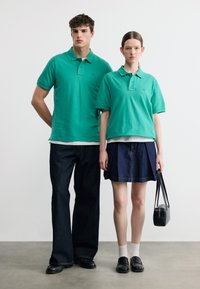 Άνδρας και γυναίκα στέκονται δίπλα-δίπλα φορώντας πράσινα πόλο μπλουζάκια· ο άνδρας με σκούρο τζιν, η γυναίκα με σκούρα φούστα κρατώντας μαύρη τσάντα χειρός.