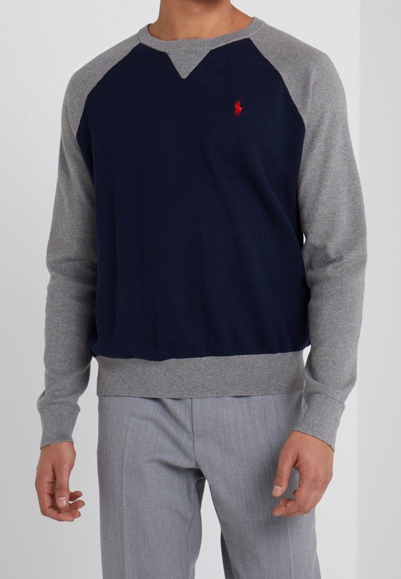 Homme portant un pull à manches longues bleu marine et gris avec un logo rouge sur la poitrine, associé à un pantalon gris clair, debout devant un fond uni.
