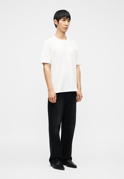 Νέος άνδρας που στέκεται φορώντας απλό λευκό μπλουζάκι, μαύρο τζιν παντελόνι με φαρδιά μπατζάκια και μ αύρα παπούτσια, σε λευκό φόντο.