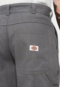 Pilkos darbo kelnės, pagamintos iš tvirto audinio, su užpakaline kišene ir stačiakampiu Dickies logotipo etikete bei siuvinėjimo detalėmis.