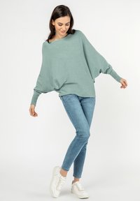 Hellgrüner Oversized-Pullover mit Fledermausärmel, kombiniert mit blauen Skinny Jeans und weißen Sneakern. Einfache, weiche Textur.