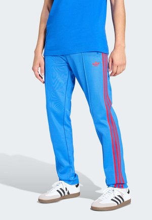 Pantalones deportivos azules con franjas laterales rojas, hechos de un material liso y ligero. Presentan un logotipo de Adidas en la cadera izquierda.