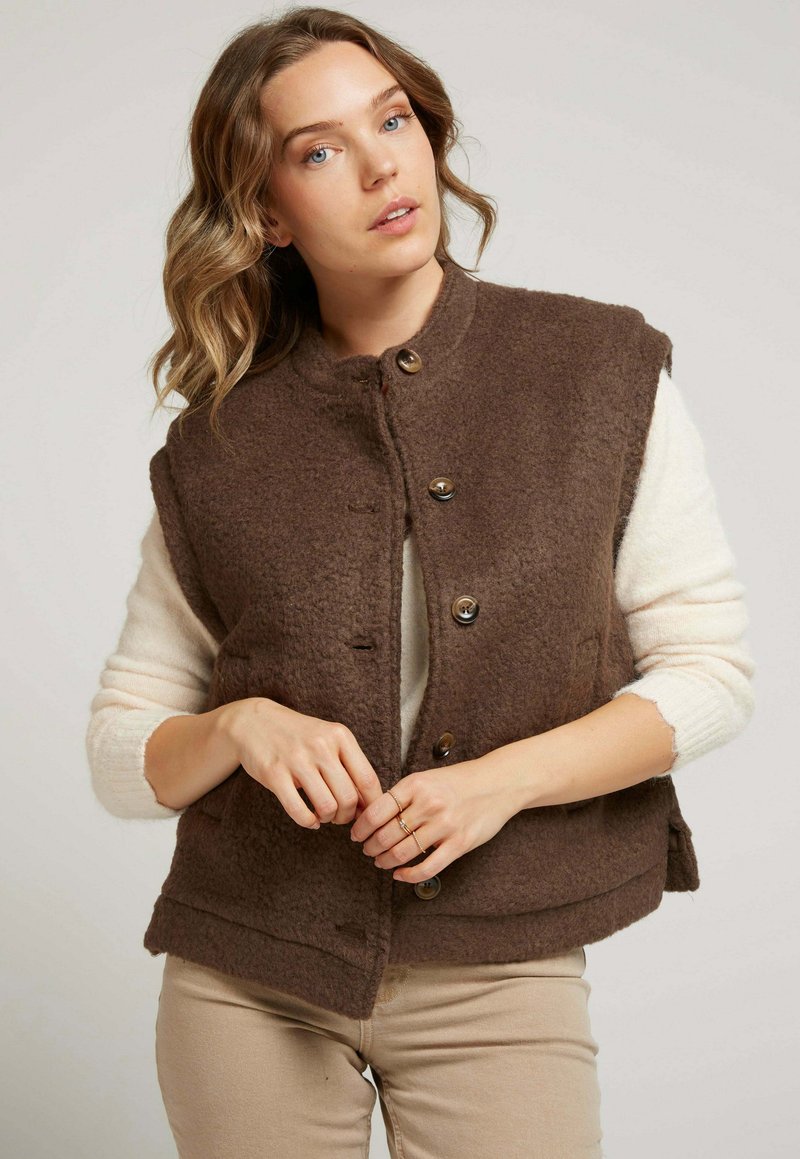 Gilet texturé marron en laine, avec un col montant, des boutons et des poches latérales, superposé à un haut à manches longues crème.