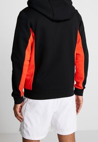 Svart hoodie med röda sidopaneler, tillverkad av mjukt material. Har en känguruficka och justerbar dragsko på huvan.