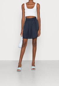 Vit cropped tank top, marinblå plisserad kjol, ljusblå väska och öppna tå-mules i blått. Släta texturer, avslappnad design och figurnära silhuett.