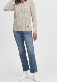 Beżowy sweter na drutach z prążkowaną strukturą, okrągłym dekoltem i małą brązową naszywką z logo. Połączony z jasnoniebieskimi dżinsami o prostych nogawkach i białymi tenisówkami.