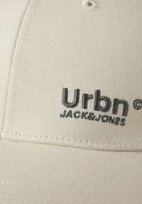 Beige baseball cap made of textured fabric, featuring an embroidered "Urbn" logo in dark gray with "JACK&JONES" underneath.