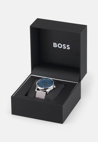 Silverfärgad klocka i rostfritt stål med en blå texturerad urtavla, omgiven av ett mesharmband, presenterad i en svart box med "BOSS"-logotyp.